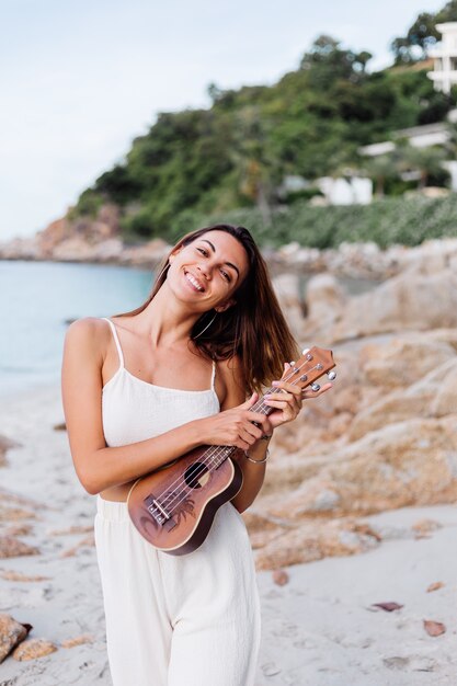 junge glückliche ruhige kaukasische Frau mit Ukulele am tropischen felsigen Strand bei Sonnenuntergang