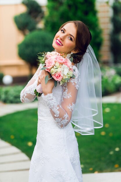 Junge glückliche schöne Braut im weißen eleganten Hochzeitskleid mit Blumenstrauß, der draußen im Park aufwirft.