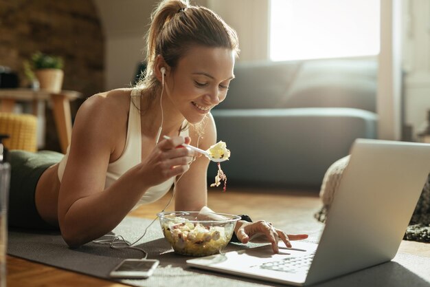 Junge glückliche Sportlerin isst Salat und benutzt Laptop, während sie sich zu Hause auf dem Boden entspannt