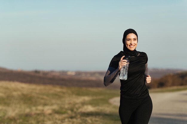 Junge glückliche sportliche Frau, die eine Flasche Wasser trägt, während sie in der Natur läuft Kopieren Sie Platz