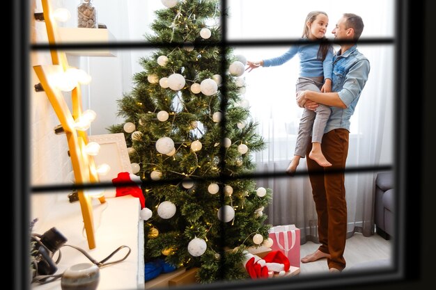 Junge große Familie feiert Weihnachten beim Abendessen, Blick von außen durch ein Fenster in ein geschmücktes Wohnzimmer mit Baum- und Kerzenlichtern, glückliche Eltern essen mit drei Kindern.