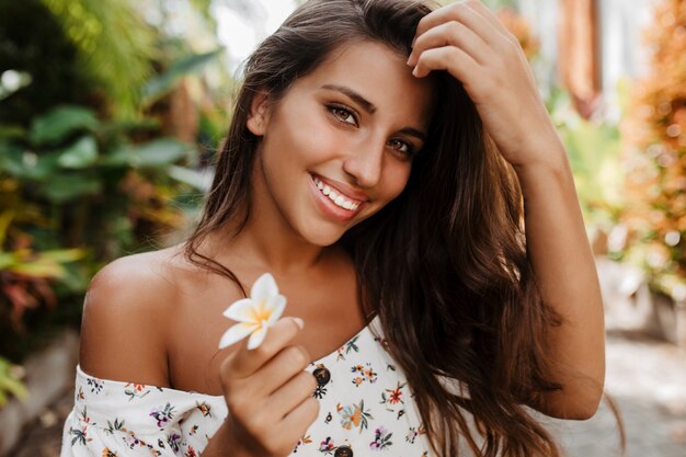 Junge grünäugige Dame lächelt und posiert mit weißer Blume im Garten