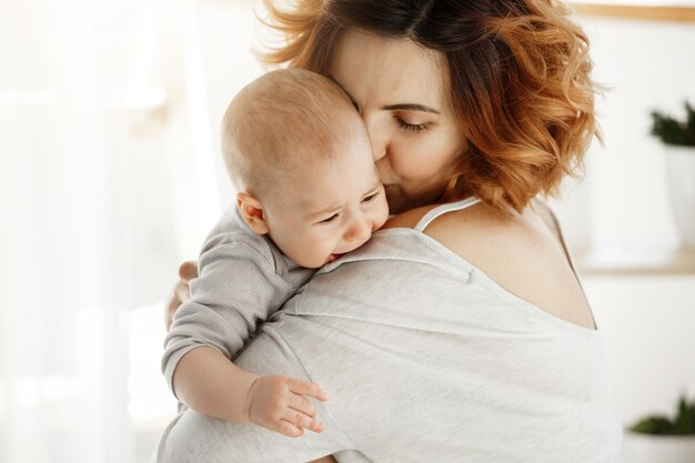 Junge gut aussehende Mutter umarmt und beruhigt ihr weinendes Baby. Kind schreit und weint auf Mutterschulter. Szene des Schutzes und der Liebe. Familienkonzept.