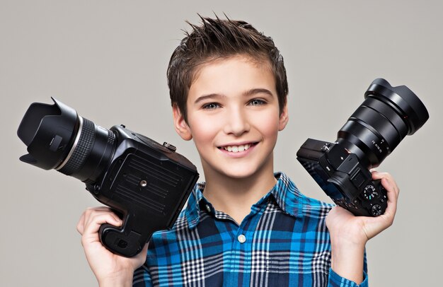 Junge hält die beiden Fotokameras. Lächelnder kaukasischer Junge mit DSLR-Kamera, die im Studio über grauem Hintergrund aufwirft