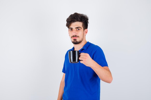 Junge hält Tasse mit der Hand im blauen T-Shirt und sieht ernst aus. Vorderansicht.