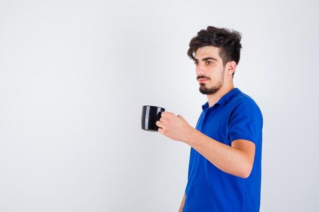 Junge hält Tasse mit der Hand im blauen T-Shirt und sieht ernst aus. Vorderansicht.