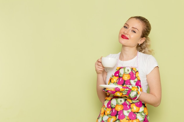 junge Hausfrau im Hemd und im farbigen Umhang, der weiße Tasse Tee hält, die auf Grün lächelt