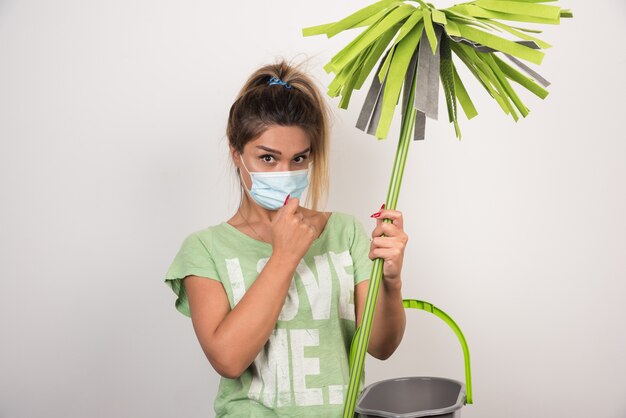 Junge Hausfrau mit Gesichtsmaske, die Mopp hält und vorne auf weiße Wand schaut.