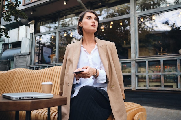 Junge, hinreißende, stilvolle Geschäftsfrau mit Handy, die während der Kaffeepause im Café auf der Straße aufmerksam wegschaut