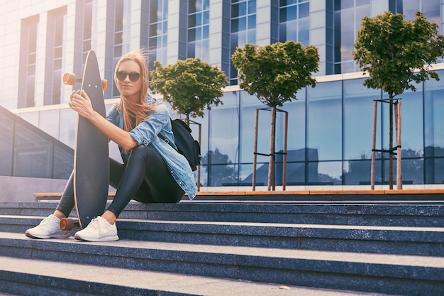 Junge Hipster-Blondine in Freizeitkleidung und Sonnenbrille, die auf Stufen gegen einen Wolkenkratzer sitzt und sich nach dem Fahren auf einem Skateboard ausruht.