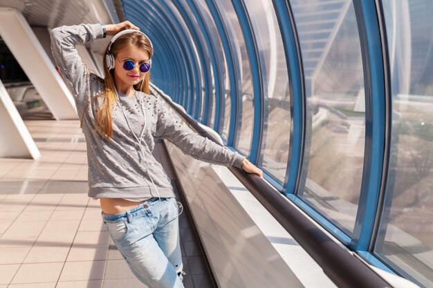 Junge Hipster-Frau, die Spaß im städtischen modernen Gebäude tanzt, gekleidet in lässigem Outfit, das Musik in Kopfhörern hört, Jeans, Pullover und Sonnenbrille tragend