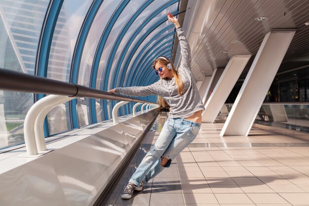 Junge Hipster-Frau, die Spaß im städtischen modernen Gebäude tanzt, gekleidet in lässigem Outfit, das Musik in Kopfhörern hört, Jeans, Pullover und Sonnenbrille tragend