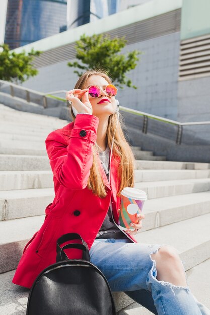 Junge Hipster-Frau im rosa Mantel, Jeans, die in der Straße mit Rucksack und Kaffee sitzen Musik auf Kopfhörern hören