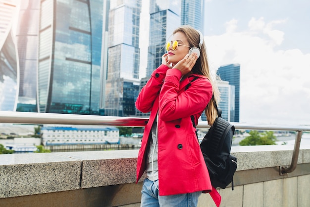 Junge Hipster-Frau im rosa Mantel, Jeans in der Straße mit Rucksack, der Musik auf Kopfhörern hört