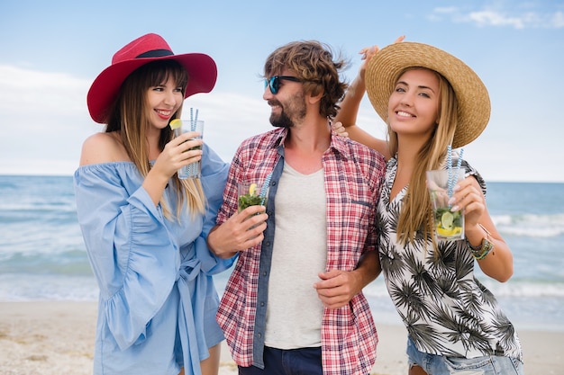 Junge hipster-gesellschaft von freunden im urlaub am strand, mojito-cocktail trinkend, glücklich positiv, sommerart, glücklich lächelnd, zwei frauen und mann, die spaß zusammen haben, reden, flirten, romantik, drei