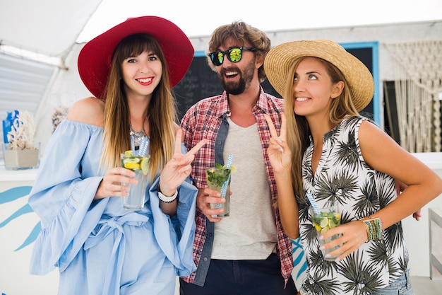 Junge Hipster-Gesellschaft von Freunden im Urlaub im Sommercafé, Mojito-Cocktails trinkend, glücklicher positiver Stil, glücklich lächelnd, zwei Frauen und Mann, die Spaß zusammen haben, reden, flirten, Romantik, drei