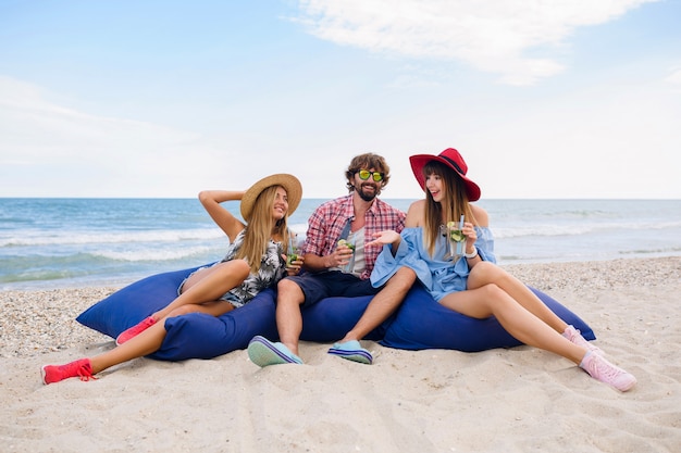 Junge Hipster-Gesellschaft von Freunden in den Sommerferien, die am Strand auf Sitzsäcken sitzen, Spaß zusammen haben, Mojito-Cocktail trinken, glücklich, lächelnd, positive, lustige Emotion, Drei-Personen-Party