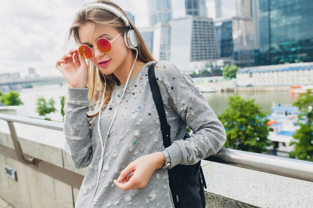 Junge Hipsterfrau, die Spaß in der Straße hat, die Musik auf Kopfhörern hört, rosa Sonnenbrille tragend