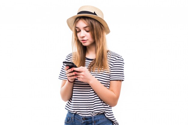 Junge Hipsterfrau im Strohhut und in der Sonnenbrille unter Verwendung des Mobiltelefons über weißer Wand