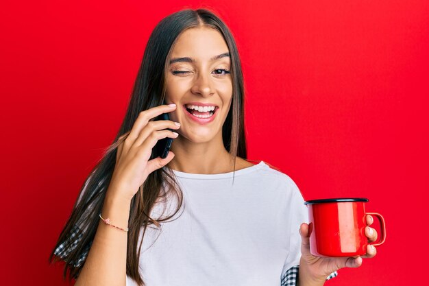Junge hispanische Frau, die auf dem Smartphone spricht und eine Tasse Kaffee trinkt, zwinkert und mit sexy Gesichtsausdruck, fröhlichem und glücklichem Gesicht in die Kamera blickt