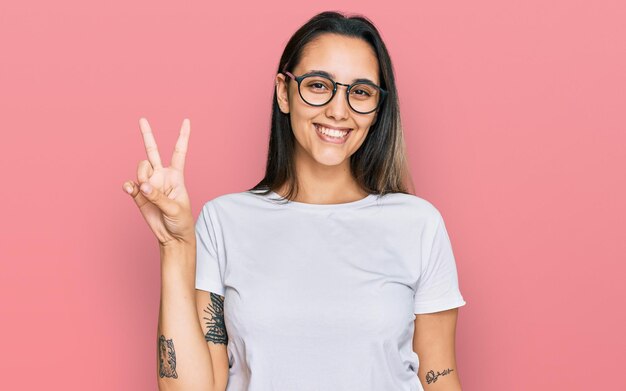 Junge hispanische Frau, die ein lässiges weißes T-Shirt trägt und lächelnd in die Kamera schaut und die Finger zeigt, die ein Victory-Zeichen machen. Nummer zwei.