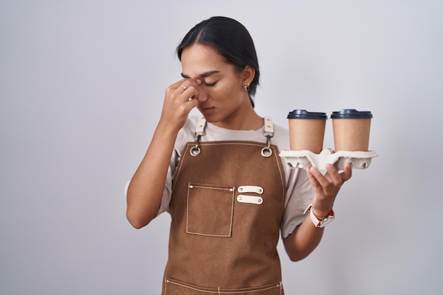 Kostenloses Foto junge hispanische frau, die eine professionelle kellnerinschürze trägt und kaffee müde hält, nase und augen reibt und müdigkeit und kopfschmerzen verspürt. stress- und frustrationskonzept.