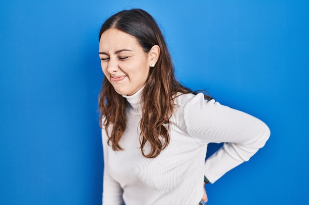 Kostenloses Foto junge hispanische frau, die über blauem hintergrund steht und unter rückenschmerzen leidet, die mit handmuskelschmerzen den rücken berühren