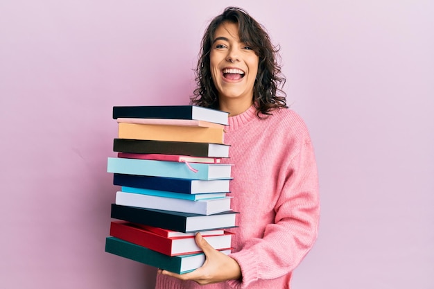 Junge hispanische Frau hält einen Stapel Bücher in der Hand, lächelt und lacht laut, weil sie einen lustigen, verrückten Witz macht