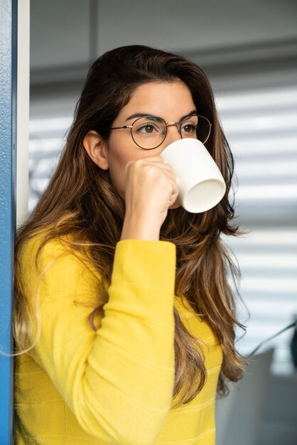 Junge hispanische Frau in einem gelben Hemd, die Kaffee trinkt und durch das Fenster schaut