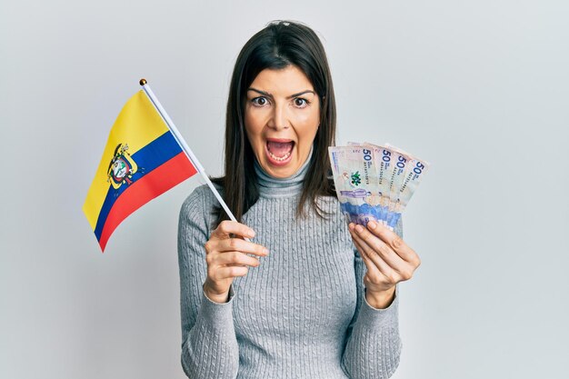 Junge hispanische Frau mit kolumbianischer Flagge und kolumbianischen Pesos-Banknoten, die verrückt und erstaunt für den Erfolg feiern, mit offenen Augen, die aufgeregt schreien.