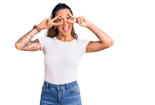 Junge hispanische Frau mit Tätowierung, die ein lässiges weißes T-Shirt trägt, das Friedenssymbol mit den Fingern über dem Gesicht macht und fröhlich lächelt und den Sieg zeigt