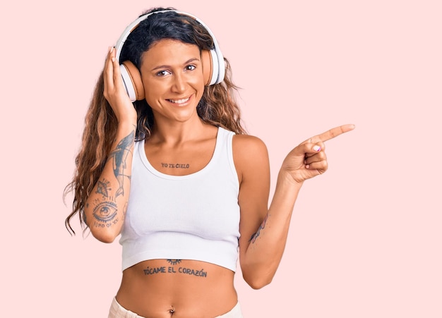 Junge hispanische Frau mit Tätowierung, die Musik über Kopfhörer hört, lächelt glücklich und zeigt mit Hand und Finger zur Seite