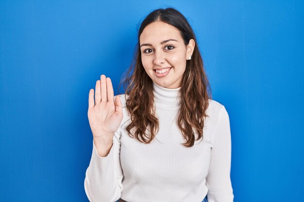 Junge hispanische Frau steht vor blauem Hintergrund und sagt „Hallo“, glücklich und lächelnd mit freundlicher Willkommensgeste