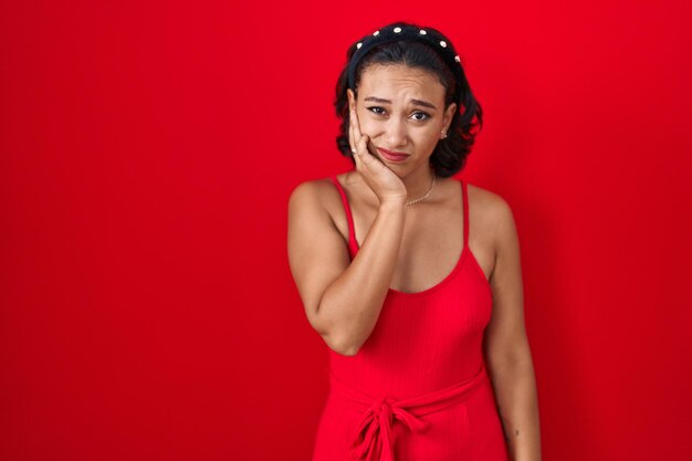 Junge hispanische Frau steht vor rotem Hintergrund und berührt den Mund mit der Hand mit schmerzhaftem Gesichtsausdruck wegen Zahnschmerzen oder Zahnerkrankungen beim Zahnarzt