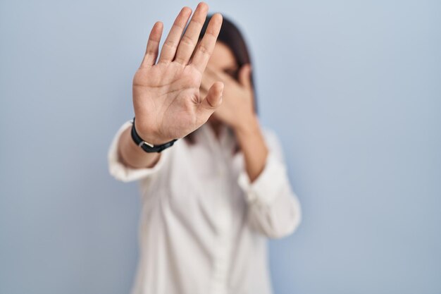 Junge hispanische Frau steht vor weißem Hintergrund und bedeckt die Augen mit den Händen und macht Stopp-Geste mit traurigem und ängstlichem Gesichtsausdruck, verlegen und negativ