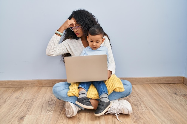 Junge hispanische Mutter und ihr Kind benutzen einen Computer-Laptop, sitzen müde auf dem Boden und reiben sich Nase und Augen, verspüren Müdigkeit und Kopfschmerzen, Stress- und Frustrationskonzept