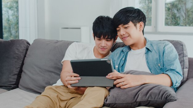 Junge homosexuelle Paare unter Verwendung der Tablette zu Hause. Das glückliche asiatische LGBTQ + -Männer entspannen sich Spaß unter Verwendung des aufpassenden Films der Technologie im Internet zusammen, während Lügensofa im Wohnzimmer.