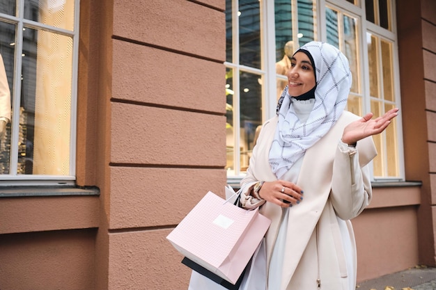 Junge hübsche arabische Frau im Hijab, die nach dem Einkaufen freudig durch die Stadtstraße geht