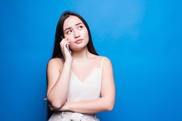 Junge hübsche asiatische Frau, die auf Handy lokalisiert über blaue Wand spricht