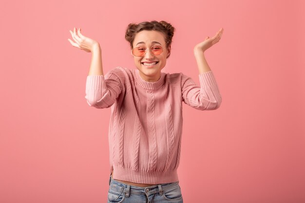 Junge hübsche aufgeregte lachende Frau mit lustigem Gesicht in rosa Pullover und Sonnenbrille im Frühlingsarttrend lokalisiert auf rosa Studiohintergrund