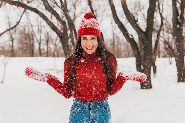 Junge hübsche aufrichtige lächelnde glückliche Frau in den roten Handschuhen und im Hut, die den gestrickten Pullover tragen, der im Park im Schnee spielt, warme Kleidung, Spaß haben
