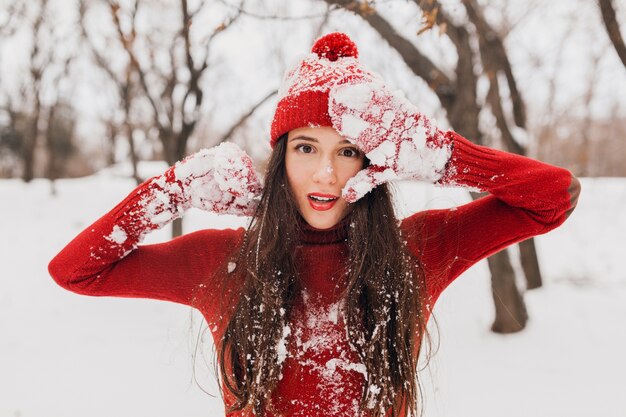 Junge hübsche aufrichtige lächelnde glückliche Frau in den roten Handschuhen und im Hut, die den gestrickten Pullover tragen, der im Park im Schnee spielt, warme Kleidung, Spaß haben