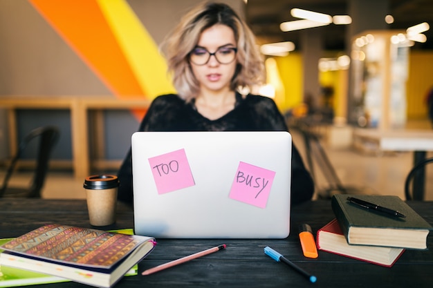 Junge hübsche beschäftigte Frau, die am Tisch sitzt und am Laptop im mitarbeitenden Büro arbeitet, Papieraufkleber, Brille tragend, Konzentration, Student im Klassenzimmer