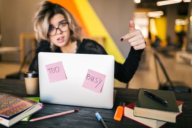 Junge hübsche beschäftigte Frau, die am Tisch sitzt und am Laptop im mitarbeitenden Büro arbeitet, Papieraufkleber, Brille tragend, Konzentration, Student im Klassenzimmer