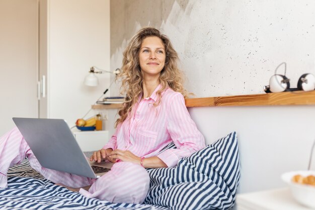 Junge hübsche blonde Frau im rosa Pyjama, der auf Bett sitzt und am Laptop arbeitet