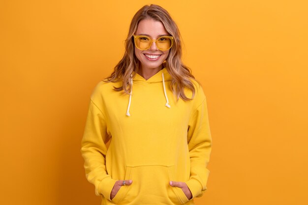 Junge hübsche blonde Frau süßer Gesichtsausdruck posiert in gelbem Hoodie auf gelber Sonnenbrille