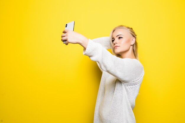 Junge hübsche Dame Mädchen im weißen Pullover macht Selfie auf ihrem Smartphone auf gelb