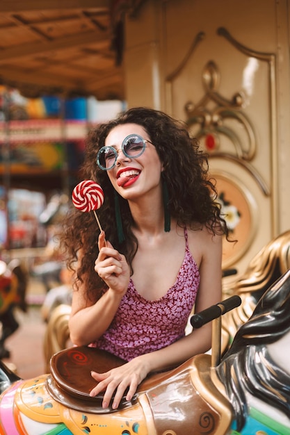 Junge hübsche Dame mit dunklem lockigem Haar in Sonnenbrille und Kleid, die mit Lolly-Pop-Süßigkeiten in der Hand steht und die Zunge in der Kamera zeigt, während sie auf Karussell im Vergnügungspark fährt