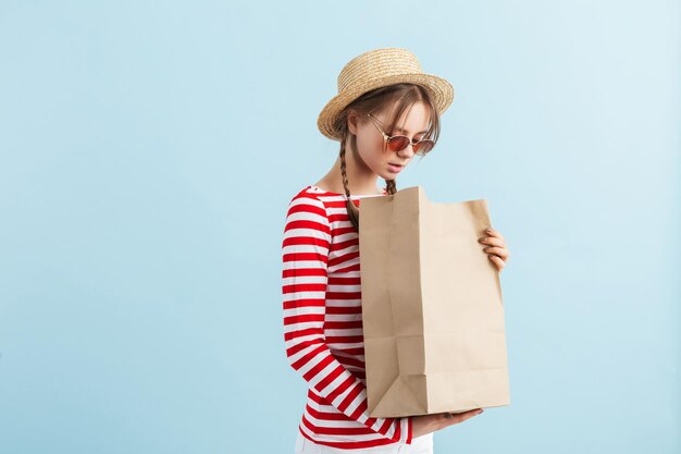 Junge hübsche Dame mit zwei Zöpfen in Strohhut und roter Sonnenbrille, die verträumt auf Papiertüte in der Hand schaut, während sie Zeit auf blauem Hintergrund verbringt