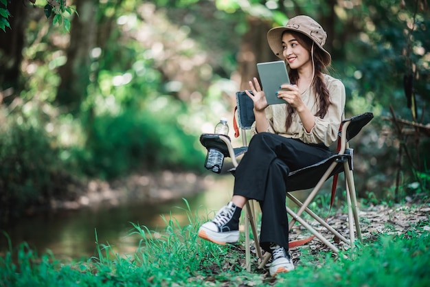 Junge hübsche Frau, die auf einem Stuhl sitzt und Tablet-Videoanrufe nutzt, während sie im Naturpark-Kopienraum campt